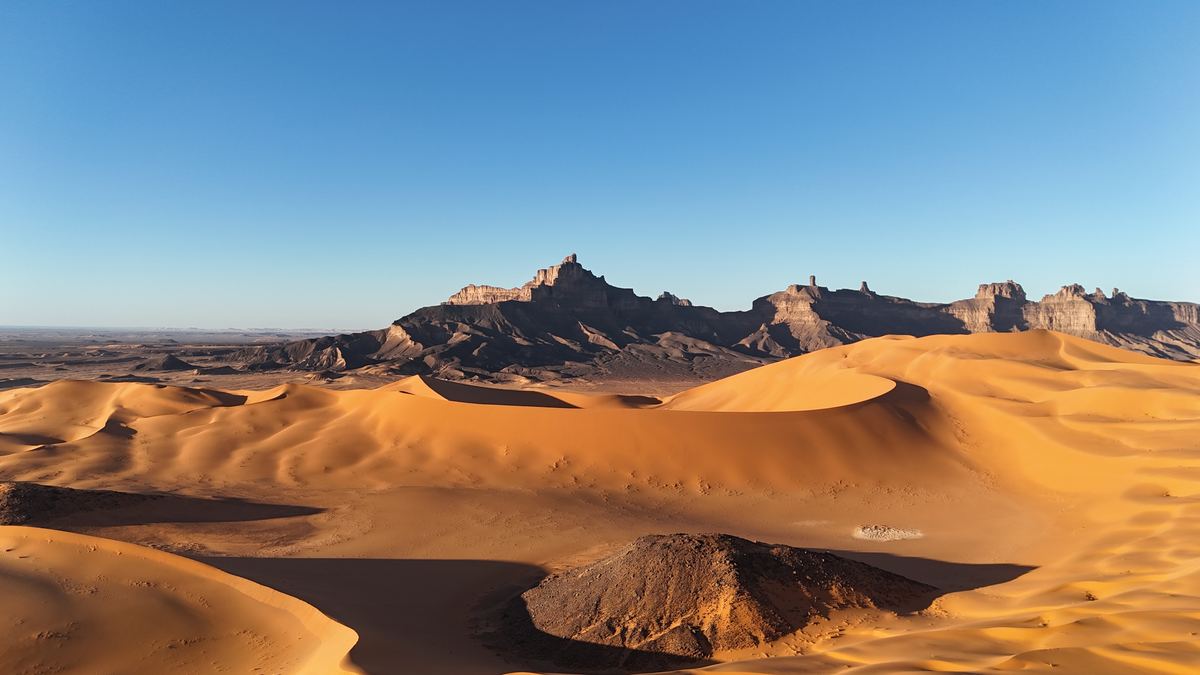 Orange dunes below dark sandstone cliffs in the Libyan Sahara